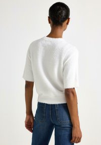 Kvinde med mørk hud og hår i en knold iført en hvid fuzzy kortærmet sweater og blå denim jeans, set fra ryggen.