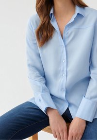 Camisa de botones azul claro con cuello clásico, mangas largas y textura suave, combinada con jeans de mezclilla oscura.