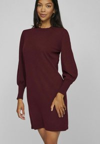 Burgundy stickad klänning med rundad halsringning, långa puffärmar, ribbade mudd och en avslappnad passform som slutar ovanför knäna. Slät yta.