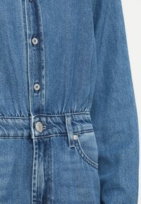 Denim jumpsuit in light blue with button closure, elastic waist, and two front pockets. Soft texture with a faded wash pattern.