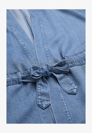 Hellblaues Denimkleidungsstück mit einer gebundenen Schleife an der Taille, das gestickte Nähte und eine weiche Stofftextur zeigt.