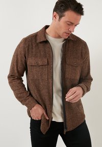 Giacca marrone in tessuto strutturato con maniche lunghe, due tasche sul petto, zip frontale e collo a camicia, indossata sopra una camicia chiara.