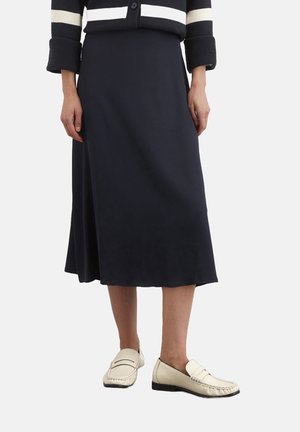 Vrouw met een marineblauwe midirok, een zwart-wit gestreepte trui en crème loafers, staand tegen een witte achtergrond.