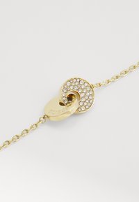 Bracciale in oro con chiusura circolare decorata con strass trasparenti, caratterizzato da una finitura lucida e una superficie liscia.