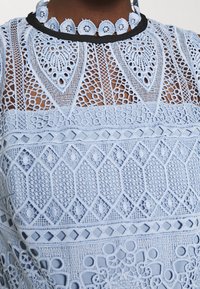 Nära bild av en ljusblå spetsplagg med intrikata blommönster och geometriska mönster samt en scalloped halsringning som är kantad med svart.