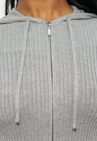 Κοντινό πλάνο ενός ατόμου που φοράει ένα ανοιχτό γκρι ριμπ ζακέτα με φερμουάρ και κορδόνια, εστιάζοντας στην περιοχή του στήθους και του λαιμού.