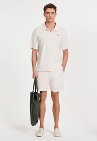 Polo shirt beige clair à manches courtes avec un logo bleu, assorti à un short. Le modèle tient un sac fourre-tout vert et porte des chaussures beiges à enfiler.