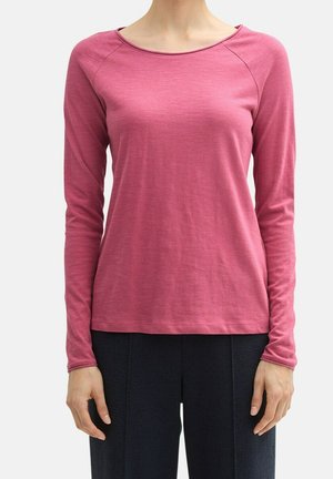 Kobieta ubrana w dopasowaną różową bluzkę z długim rękawem i okrągłym dekoltem oraz czarne spodnie, stojąca na tle jednolitego jasnego tła.