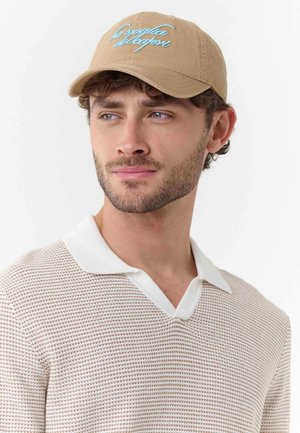 Man with light beard wearing beige cap with blue script and white-collared textured beige sweater, looking slightly to the side.