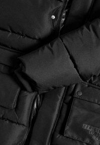 Zwarte gewatteerde jas met een dikke, doorgestikte ontwerp. Ritsluiting, drukknoopsluiting en een patchzak met tekstdetail. Zachte textuur.