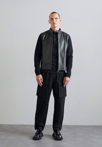 Black leather vest over a black long-sleeve shirt, paired with black cargo pants. Features a zip closure and large pockets. Black shoes.