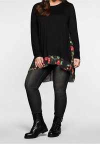 Zwarte lange mouw top met een bloemenrand, gecombineerd met donkergrijze skinny jeans en zwart veterschoenen. De rand heeft rode en groene bloemenpatronen.