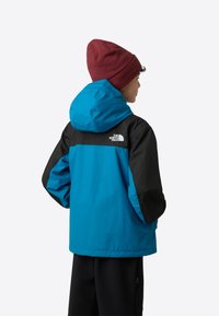 Blauwe en zwarte waterdichte jas met capuchon, voorzien van elastische manchetten en een logo op de bovenrug. Draagt een bordeauxrode beanie.