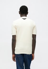  Μπλούζα πόλο σε κρεμ χρώμα με κοντά μανίκια και υφή υφάσματος, με αντίθετη μαύρη λεπτομέρεια στον πίσω γιακά και ριμπ τελειώματα στα μανίκια.