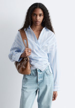 Mladá žena s dlhými kučeravými vlasmi, ktorá nosí svetlomodrú zavinovaciu blúzku a svetlé džínsové nohavice, drží hnedú koženú kabelku na ramene.