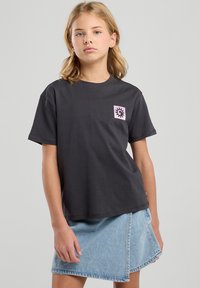 Mustat puuvillapaita, jossa on laatikkomainen malli, lyhyet hihat ja vaaleanpunainen graafinen logo rinnassa, yhdistettynä vaaleansiniseen farkuhameeseen.