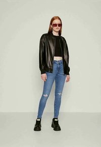 Jovem mulher vestindo um top cropped preto, uma jaqueta de couro preta, jeans skinny azuis rasgados, botas pretas robustas e óculos de sol vermelhos oversized.