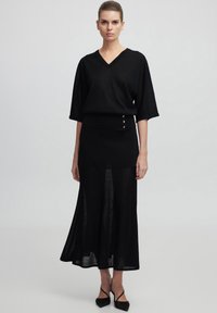 Haut en maille noir avec un col en V et des manches larges, associé à une jupe longue semi-transparente noire avec des accents de boutons dorés.