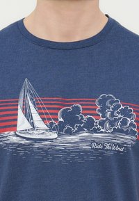 Ιστιοφόρο με γεμάτα πανιά στο νερό κοντά σε στυλιζαρισμένα σύννεφα και κόκκινες οριζόντιες γραμμές, με το κείμενο «Ride The Wind» σε μπλε ύφασμα.