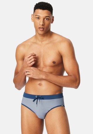 Jonge man met kort krullend haar, gekleed in een blauwe zwemslip met patroon, staand tegen een effen lichte achtergrond, zijn handen verstelend.