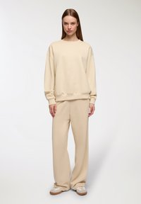 Felpa in cotone beige e pantaloni abbinati con una vestibilità rilassata, con polsini a coste e una texture liscia. Sneakers bianche con suole marroni.