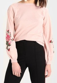 Hellrosa gestrickter Pullover mit Blumenstickerei an den Ärmeln. Verfügt über einen Bootsausschnitt und Ballonärmel. Kombiniert mit schwarzen Hosen.