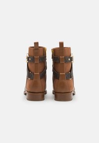Bottes chevilles marron avec une finition en cuir lisse, deux sangles sombres avec boucles dorées, et un talon en bois empilé. Accents en tissu texturé.