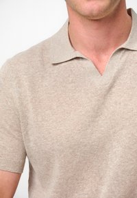 Beige knitted polo shirt with a short sleeve, V-neck design, featuring a subtle textured pattern and ribbed cuffs.