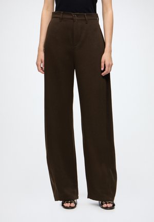 Person trägt hochgeschnittene, weit geschnittene dunkelbraune Hose und schwarze Sandalen mit offenen Zehen und goldener Kettendetail, steht vor weißem Hintergrund.