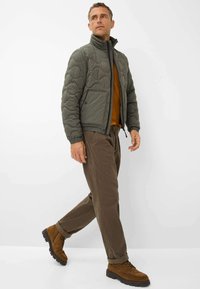 Gevoerde olijfgroene jas met een hoge kraag, ritssluiting en twee zijzakken, gedragen over een bruine shirt en gecombineerd met een bruine broek en laarzen.