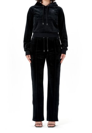 Vrouw gekleed in een zwarte velours hoodie met trekkoorden, bijpassende hoge taille broek en zwarte open-toe hakken, staand tegen een witte achtergrond.