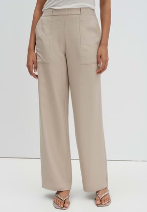 Beige wijde broek met voorzakken, gedragen met een wit topje en sandaaltjes met open teen op een effen lichte achtergrond.