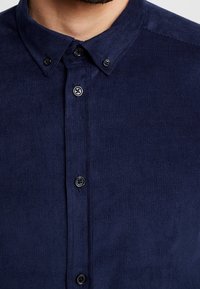 Laivastonsininen ribbineulospaita, jossa on nappikiinnitteinen kaulus, mustat napit ja poikkiharmaa tekstuuri. Muotoilu korostaa räätälöityä istuvuutta ja toiminnallisia yksityiskohtia.