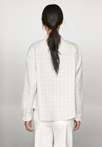 Chemise à carreaux gris clair en tissu doux, avec une coupe décontractée, des manches longues et un design à empiècement au dos. Associée à un pantalon blanc.