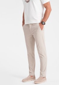 Mężczyzna w beżowych, dopasowanych spodniach, jasnobeżowych butach typu slip-on, białej koszulce z grafiką i czarnej nawijanej zegarku, stoi z rękami w kieszeniach.