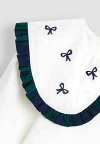Weißer Stoffdetail mit marineblauen gestickten Schleifen und einer Volantkante mit grün-blauem Karomuster.