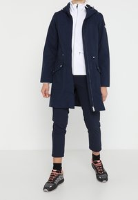 Marinfärgad vattentät parka med huva, framdragkedja och två fickor. Bärs över en vit jacka med dragkedja och marinblå byxor, i kombination med sportiga svarta skor.