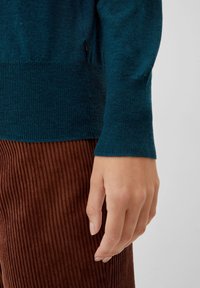 Pull en tricot bleu canard avec des poignets côtelés, associé à un pantalon en velours côtelé marron. Gros plan sur une main reposant contre un tissu texturé.
