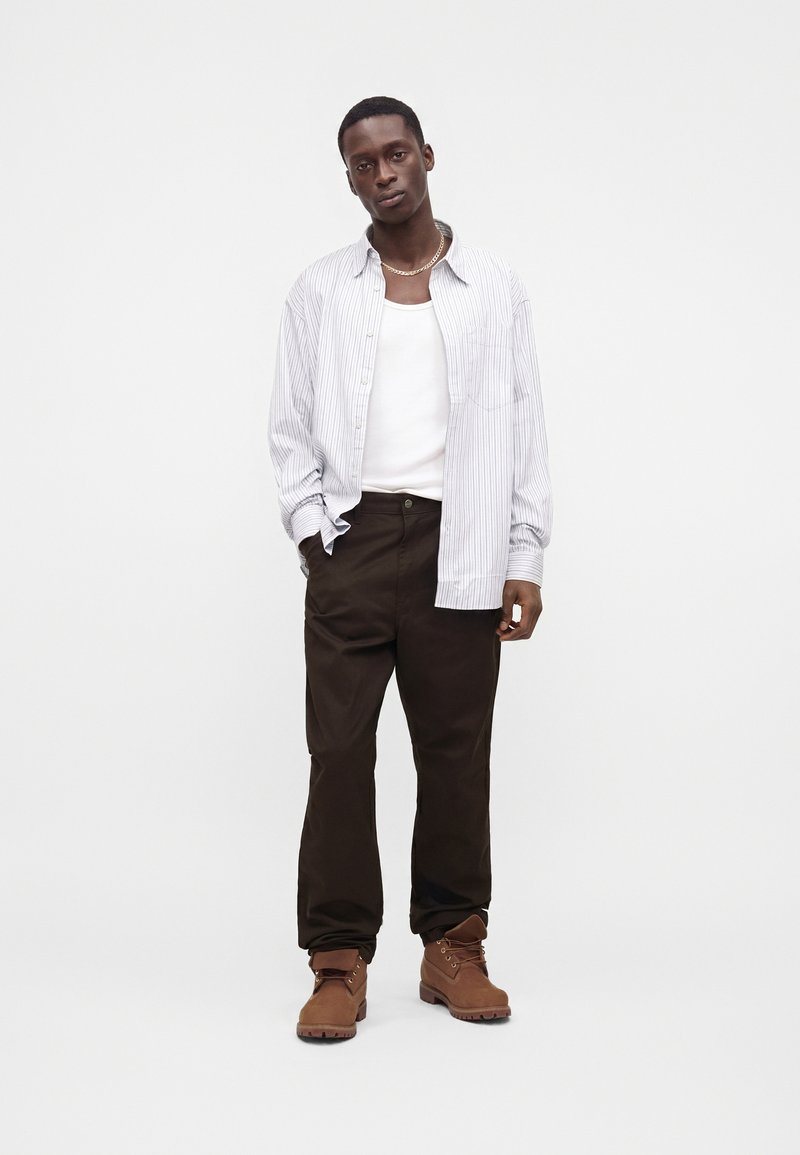 Homme portant un pantalon marron, des bottes couleur beige, un débardeur blanc et une chemise blanche à rayures ample, debout sur un fond blanc.