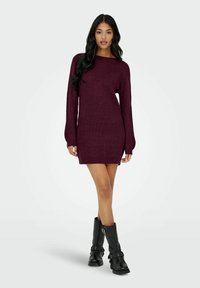 Donna che indossa un vestito a maglia a maniche lunghe di colore viola scuro e stivali neri fino a metà polpaccio, in piedi contro uno sfondo chiaro e uniforme.