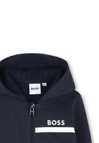 Sweat à capuche bleu marine avec fermeture éclair devant, logo blanc "BOSS" et une bande horizontale blanche sur la poitrine.