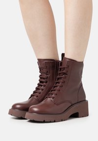 Hnědé kožené kotníkové boty s kulatou špičkou, šněrováním a bočním zipem. Obsahují robustní platformovou podrážku s protiskluzovým vzorem.