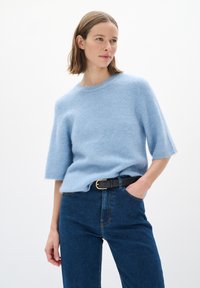 Ljusblå, kortärmad, fluffig stickad tröja med rund halsringning, bärs tillsammans med mörk denimjeans och ett svart bälte med ett guldspänne.