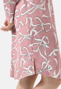 Rosa tygklänning med ett upprepat mönster av vita rosetter, långa ärmar och en sidoklyvning för rörlighet. Slät textur, avslappnad design.