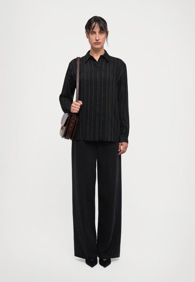 Chemise noire à rayures verticales avec des manches longues, devant à boutons, associée à un pantalon noir ample et un sac bandoulière marron.