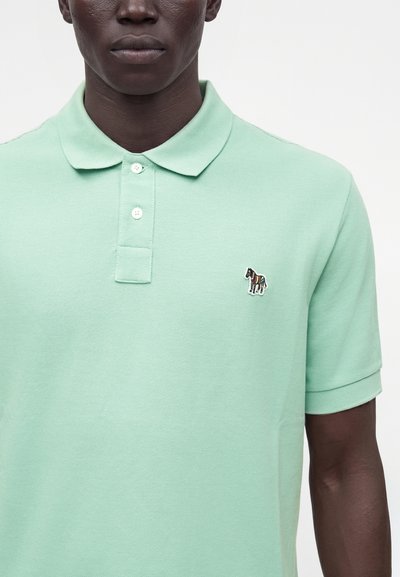 Homme portant un polo vert clair avec un petit logo zèbre coloré sur la poitrine gauche, deux boutons fermés, manches courtes.