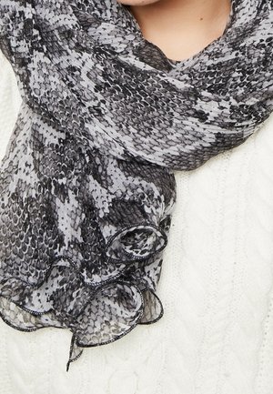 Personne portant une écharpe noire et grise à motif peau de serpent sur un pull blanc en maille torsadée, montrant le cou et la partie supérieure de la poitrine.