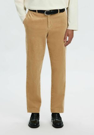 Homme portant un pantalon en velours côtelé beige avec une ceinture tissée noire, un haut crème à manches longues et des mocassins en cuir noirs, debout sur fond blanc.