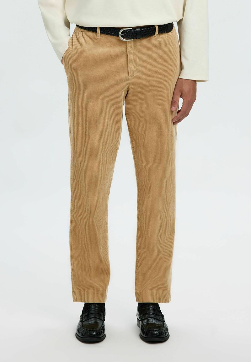 Homme portant un pantalon en velours côtelé beige avec une ceinture tissée noire, un haut crème à manches longues et des mocassins en cuir noirs, debout sur fond blanc.