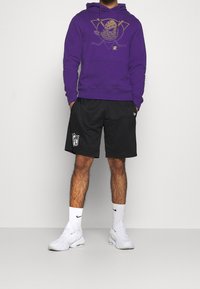 Sudadera púrpura con un diseño gráfico y pantalones cortos de malla negros con un logo de la NFL. Combinado con zapatillas blancas y calcetines.